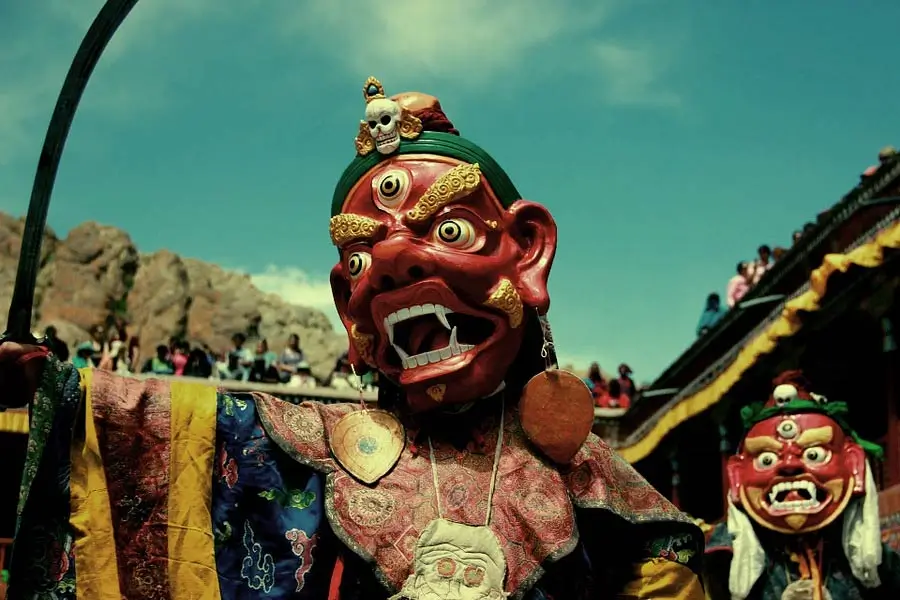 Red demon mask performer in colorful robes during a festival; crowd and temple rooftops in the background.