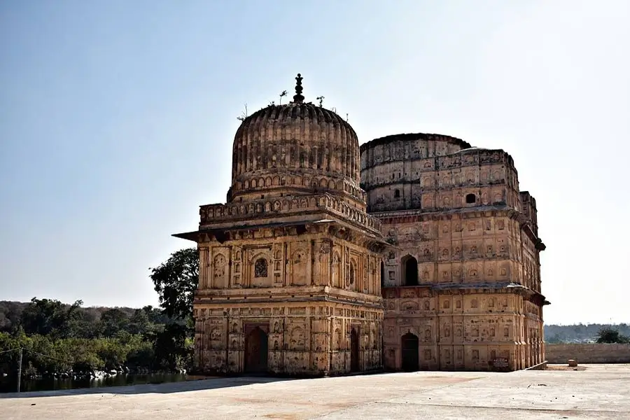Two ornate sandstone mausoleums with domed roofs and carved panels on a sunny open plaza in India.